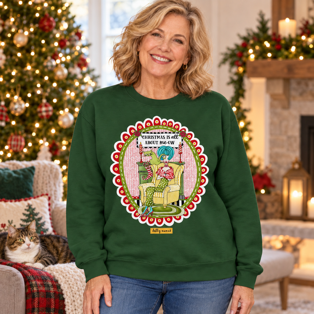 Woman wearing a forest green “Christmas Is All About me-ow” sweatshirt in a Christmas living room with a cat resting nearby.