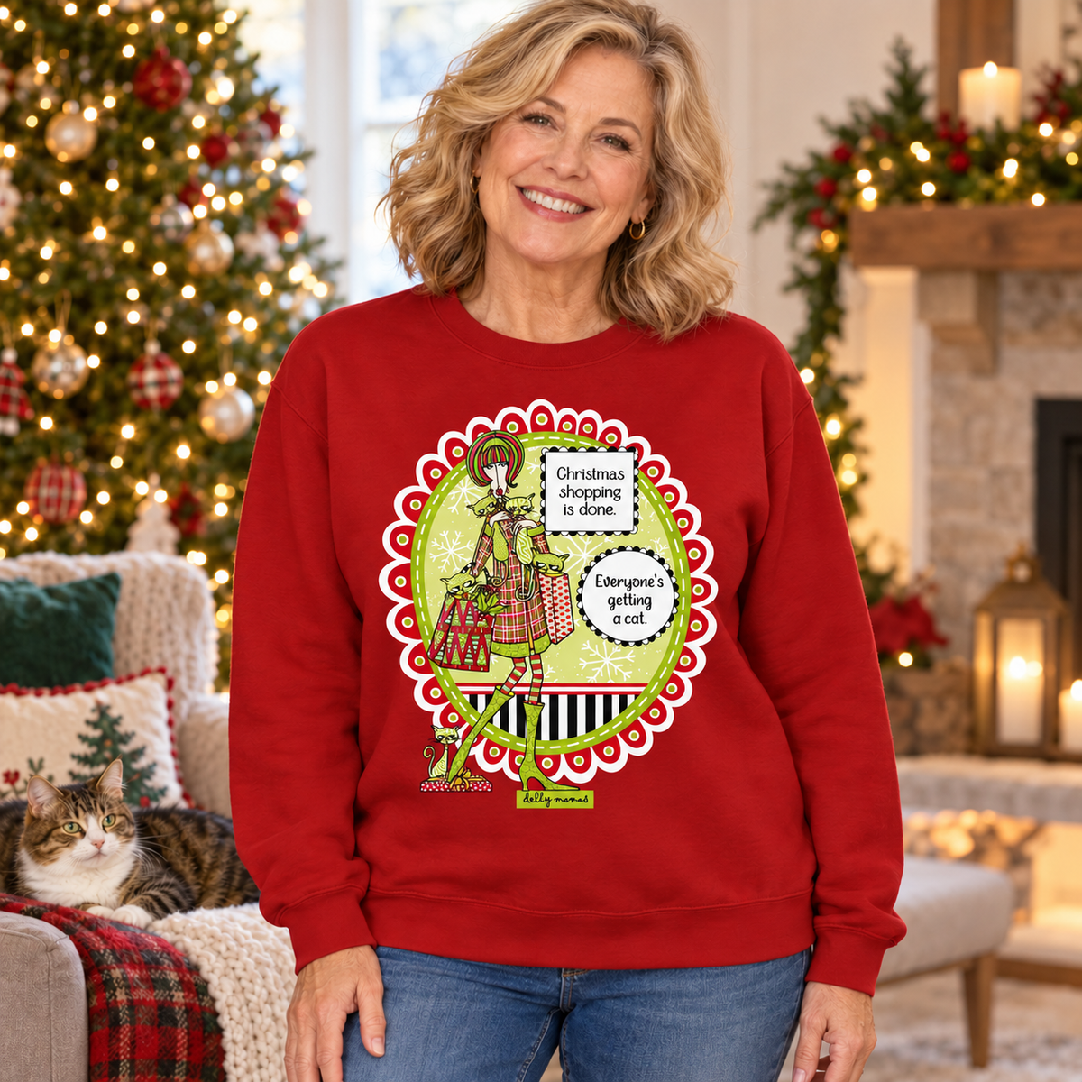 Woman wearing a cardinal red Christmas Shopping Is Done sweatshirt beside a Christmas tree, fireplace, and lounging cat.