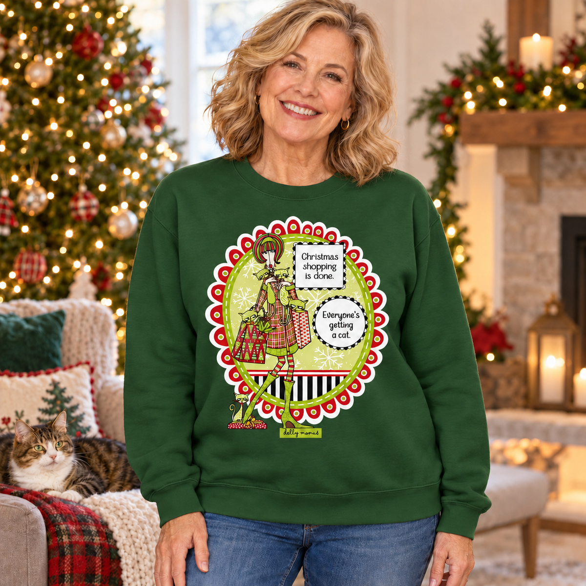 Woman wearing a forest green Christmas Shopping Is Done sweatshirt in a Christmas living room with a cat resting nearby.