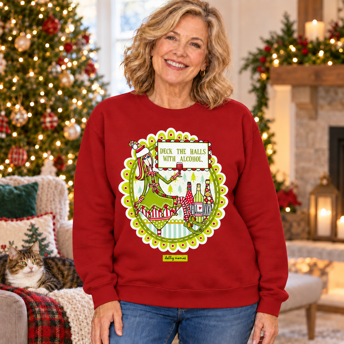 Woman wearing a cardinal red Dolly Mamas “Deck the Halls with Alcohol” sweatshirt beside a Christmas tree, fireplace, and lounging cat.