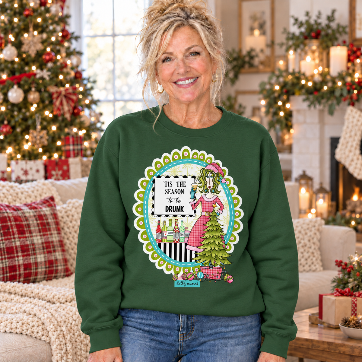 Woman wearing a forest green Dolly Mamas ’Tis the Season to Be Drunk sweatshirt beside a decorated Christmas tree and festive fireplace.
