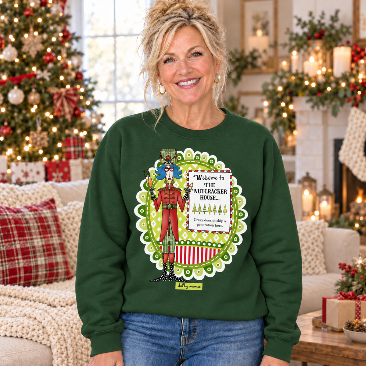 Woman wearing a forest green Dolly Mamas Welcome to the Nutcracker House sweatshirt beside a decorated Christmas tree and festive fireplace.