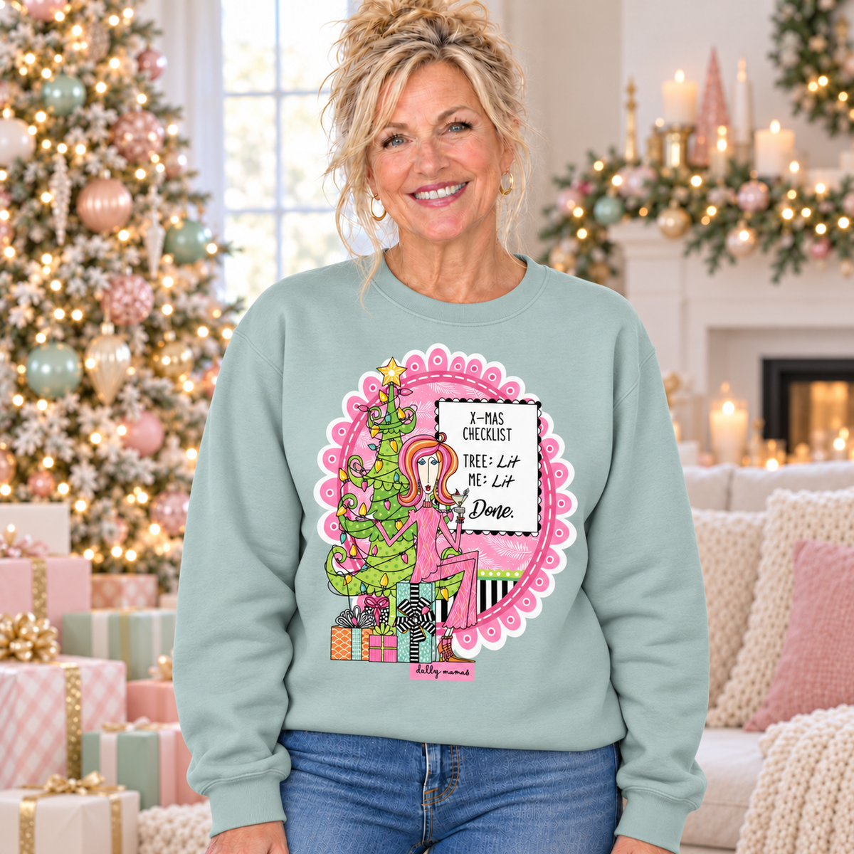Woman wearing an agave Dolly Mamas X-mas Checklist sweatshirt beside a Christmas tree with pastel ornaments and wrapped gifts.