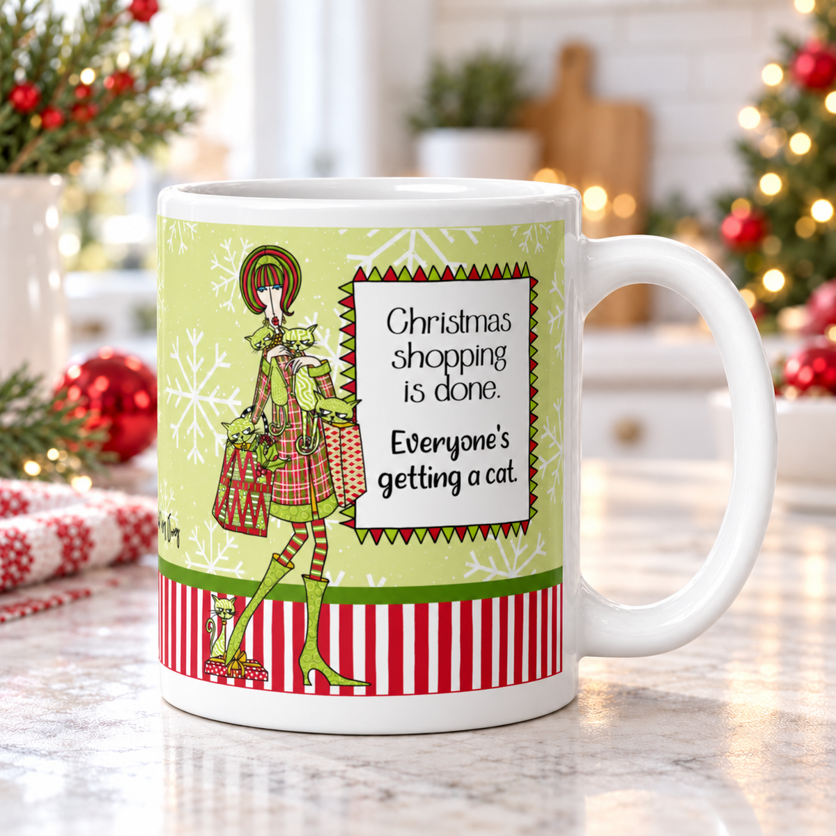 Dolly Mama “Christmas Shopping Is Done. Everyone’s Getting a Cat” mug on a kitchen counter, handle right, with Christmas greenery and red ornaments.