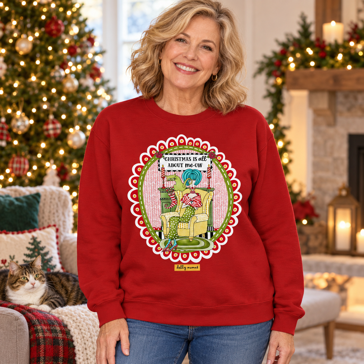  Woman wearing a cardinal red “Christmas Is All About me-ow” sweatshirt beside a Christmas tree and a lounging cat.
