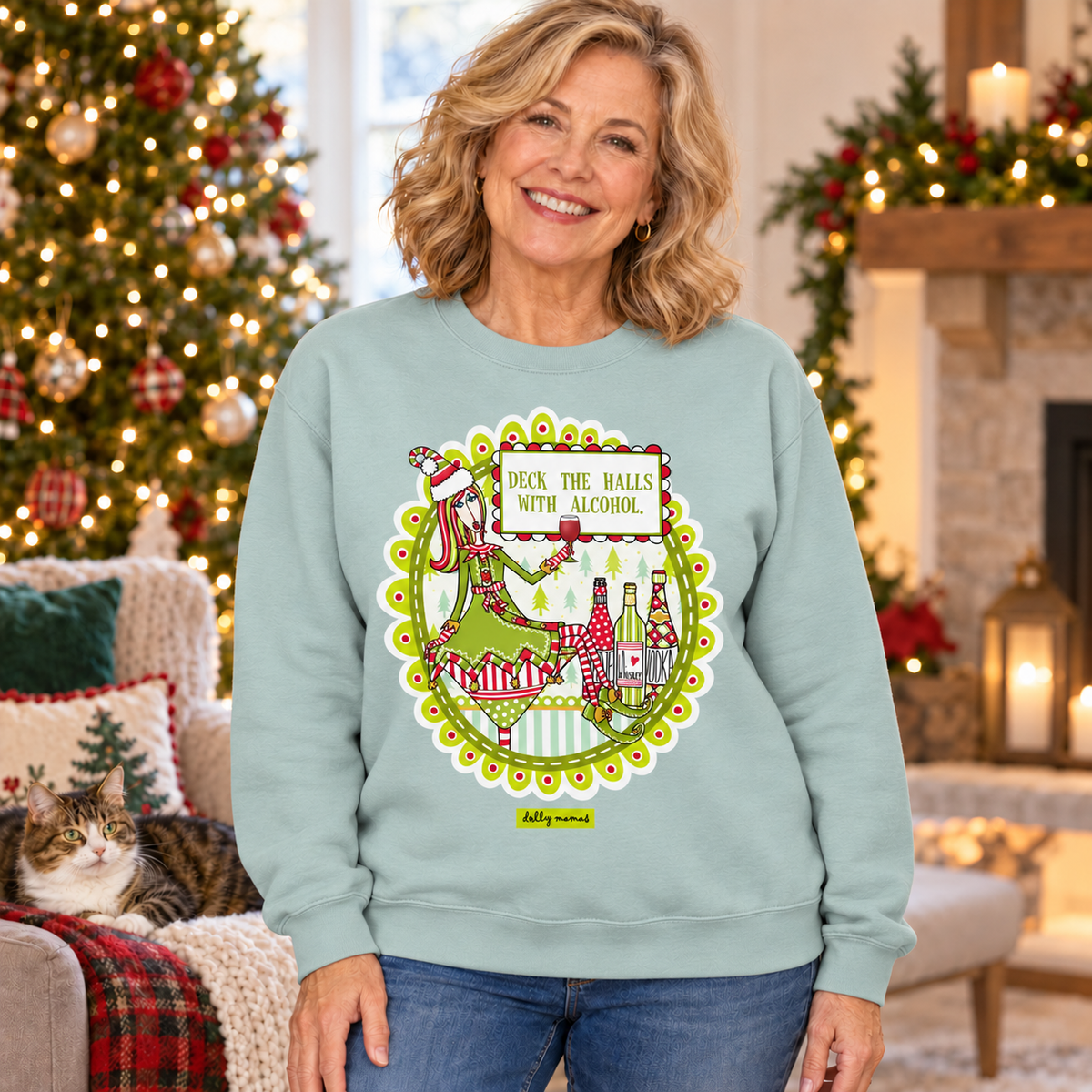 
Woman wearing an agave green Dolly Mamas “Deck the Halls with Alcohol” sweatshirt beside a Christmas tree, fireplace, and lounging cat.
