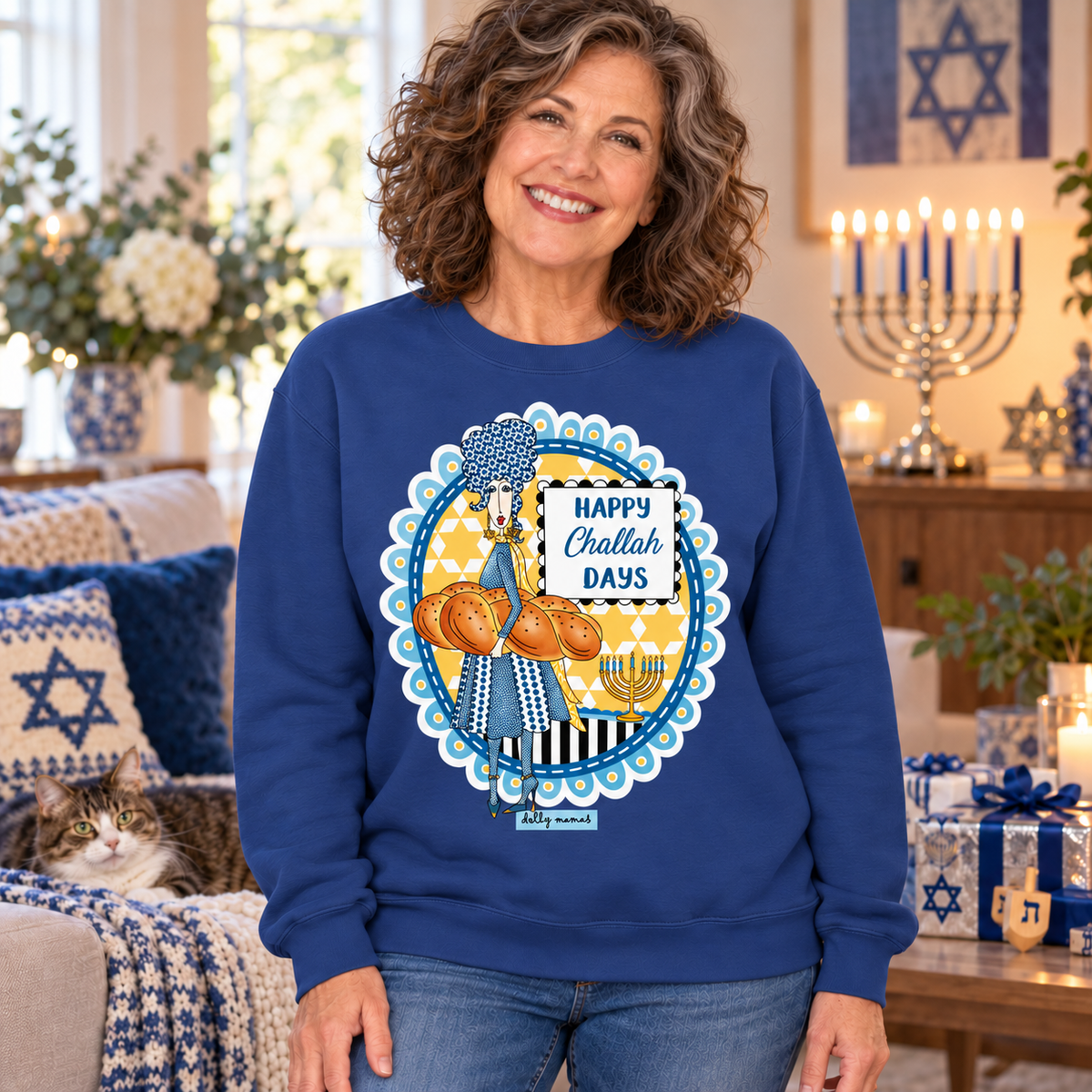 
Woman wearing a royal blue Dolly Mamas “Happy Challah Days” sweatshirt beside a lit menorah, wrapped gifts, and a lounging cat.
