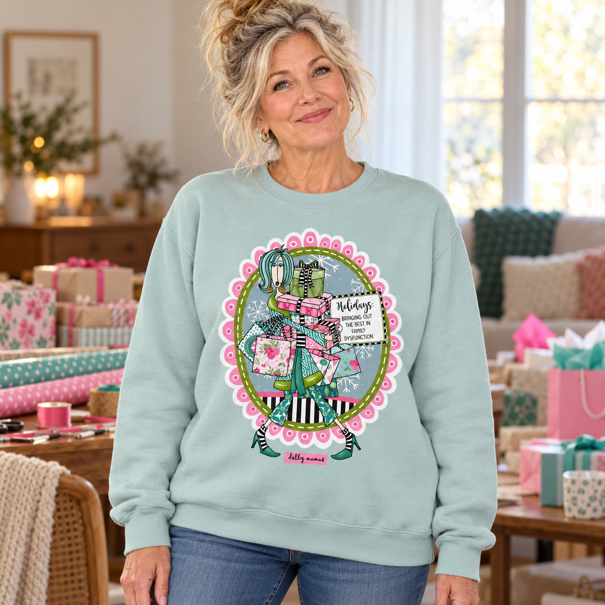 Woman wearing an agave green Dolly Mamas holiday sweatshirt beside wrapped gifts and colorful gift-wrapping supplies.
