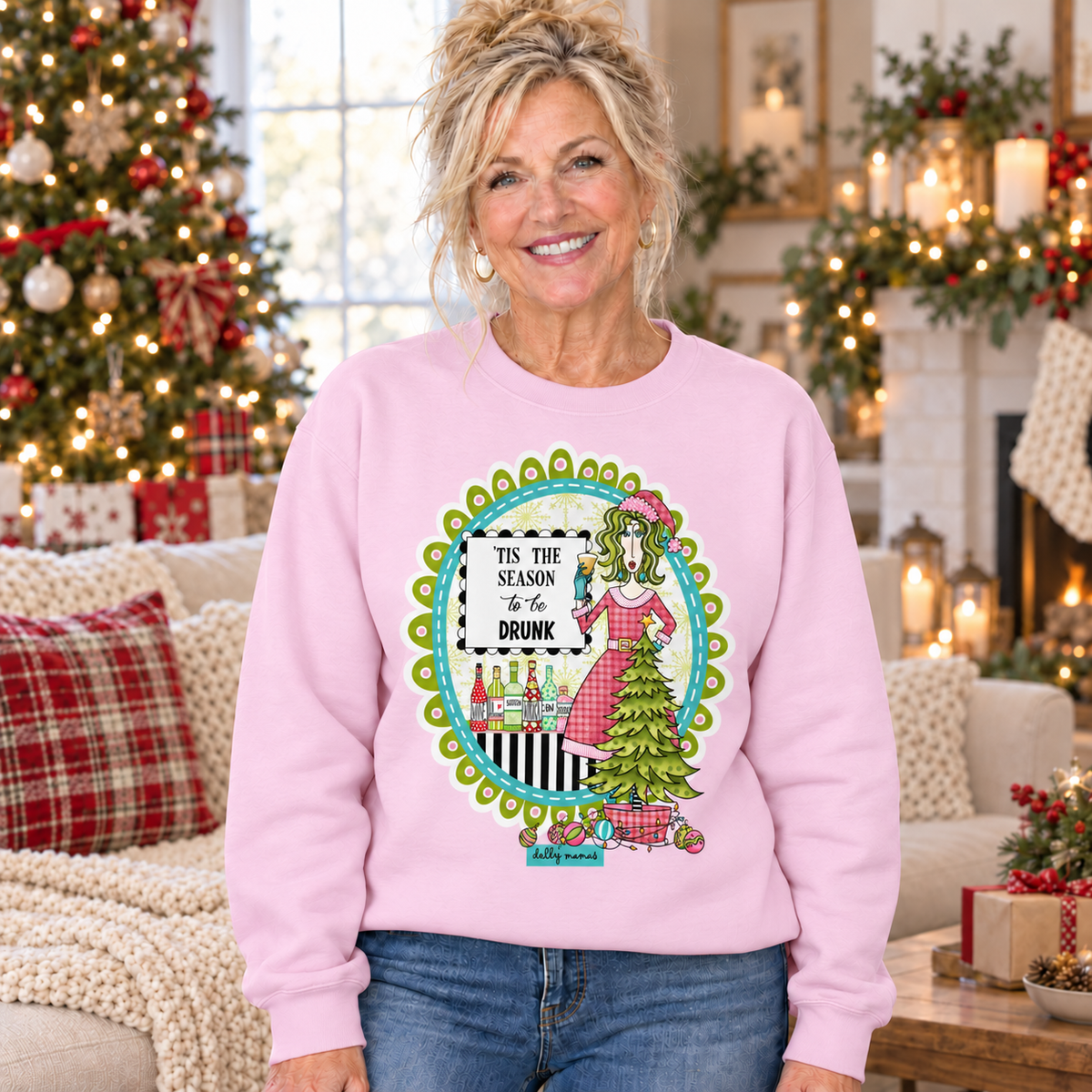 Woman wearing a pale pink Dolly Mamas ’Tis the Season to Be Drunk sweatshirt beside a decorated Christmas tree and festive fireplace.
